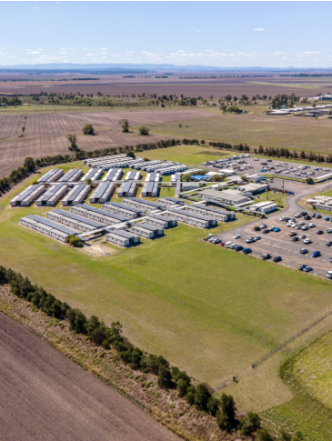 a wide aerial photograph of a rural suburban area. there are several buildings and structures scattered across a large grassy field with a few scattered trees, including a large parking lot filled with cars. the buildings look like small sheds with a row of them facing towards the camera. the ground is mostly grass and the sky is clear with very light wispy clouds in the distance.