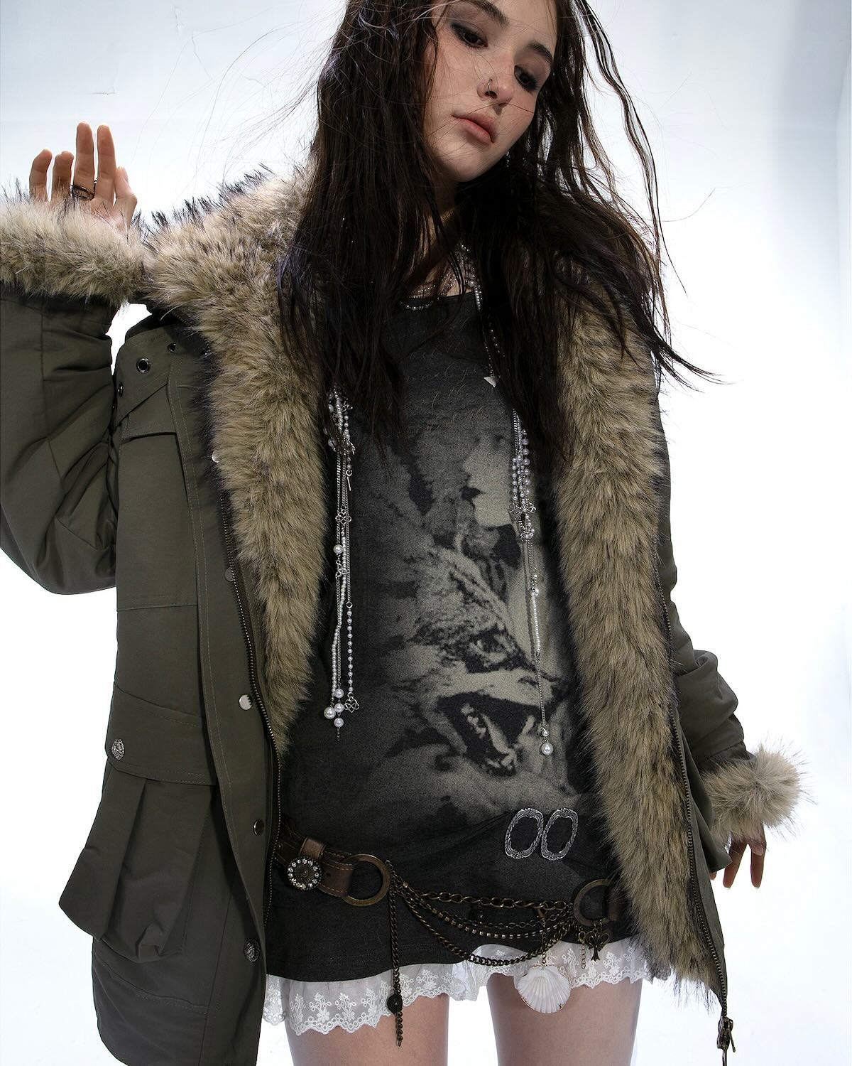 a young woman with long dark hair models a stylish olivegreen parka with a thick faux fur trim.  the parka is worn over a dark grey graphic tshirt and a white lace skirt. she is accessorized with multiple necklaces, chains and rings. the overall style is edgy and bohemian. the photo is a studio shot against a white background.