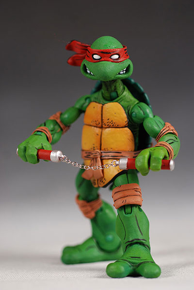 a teenage mutant ninja turtle action figure, specifically raphael, is posed holding nunchucks. he is green with a red bandana and an orange shell. he has brown wrappings on his wrists and knees. the figure is standing on a gray surface against a gray background.