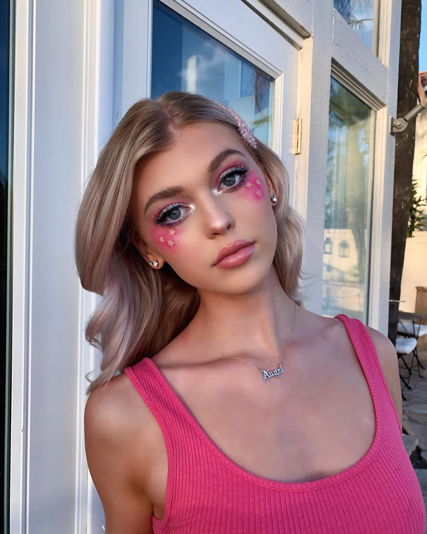 a portrait of loren gray, a young woman with blonde hair styled in loose waves. she has elaborate makeup with pink stars on her cheeks and sparkling eyeshadow. she wears a pink tank top and a necklace with the word "angel". a sparkly hair clip adorns her hair. she is positioned in front of a door and window with outdoor scenery visible in the background.