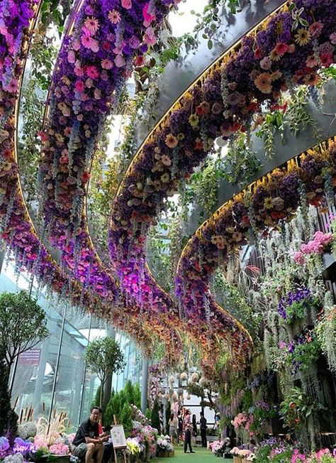 the image showcases a stunning floral installation, a breathtaking display of vibrant purple, pink, and other colorful flowers hanging from the ceiling in elegant arches. these arches create a beautiful walkway leading through a lush garden setting. the flowers are densely packed creating a visually impressive effect. a man is seen sitting in the garden near the walkway, observing the floral spectacle. the overall scene is a blend of both indoor and outdoor elements, highlighting the beauty of nature combined with artistic design. the environment appears to be a public place, possibly a garden or indoor public space, maybe even a temporary installation for an event.