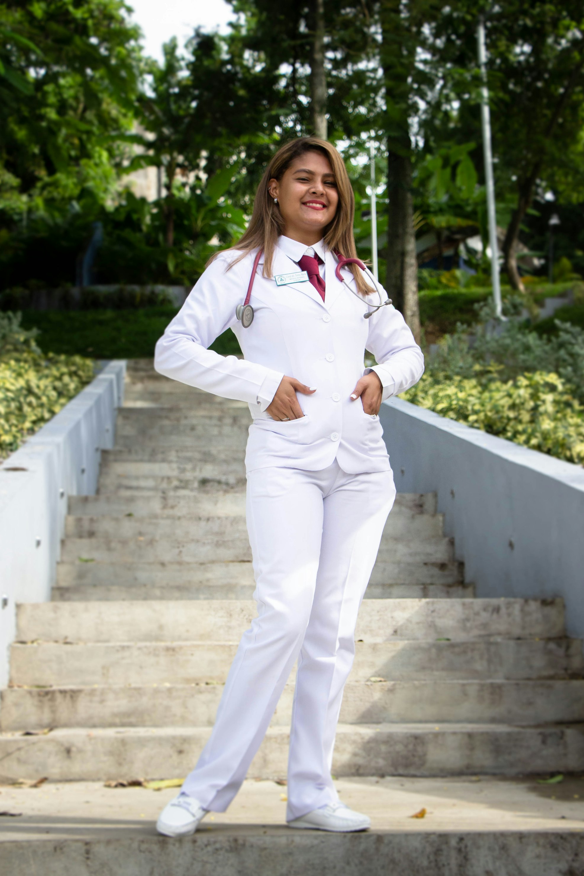 a smiling young woman, likely a medical student or recent graduate, poses confidently on a set of stairs in a park. she is dressed in a crisp white pantsuit and lab coat, with a stethoscope around her neck. her attire and posture project professionalism and success in the medical field.  the background features lush green trees and landscaping, providing a pleasant, outdoor setting for the portrait.  the overall mood is one of positivity and achievement.