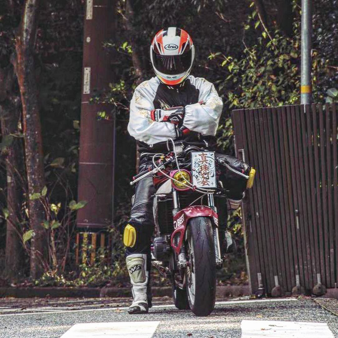a motorcyclist in full gear, helmet and all, stands next to his motorcycle in a crosswalk. the motorcycle has a red and black body and a white stripe on the front tire. the rider is wearing a white jacket with black accents and black pants. the background is a blurred out street, trees, and a wooden fence.