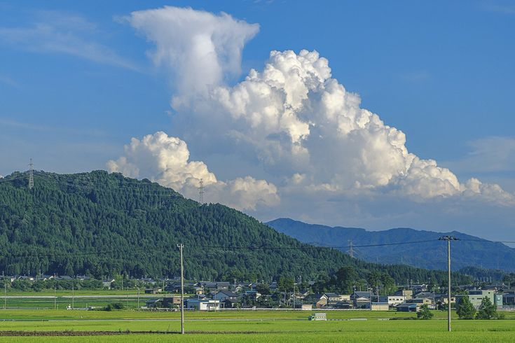 a landscape featuring a green field in the foreground, a village nestled at the foot of a lush green mountain, and expansive clouds in a blue sky. electricity pylons dot the landscape.