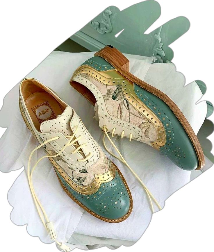 a closeup shot of a pair of women's floral patterned oxford shoes. the shoes are predominantly beige and teal with gold accents. they have a wingtip design and are made of leather. the shoes are positioned on a white surface, and the background is a blurry image of a light teal colored surface. the image is styled in a flatlay fashion, and the shoes are the main focus. the overall aesthetic is vintage and elegant.