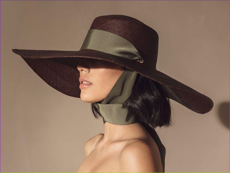 a woman wearing a widebrimmed brown hat with a green ribbon. the hat is sitting low on her head, and the ribbon is tied around her neck. she is looking to the right. her hair is black and pulled back and there are no other accessories on her. she is wearing a neutral colored outfit.  she is standing in a studio setting.