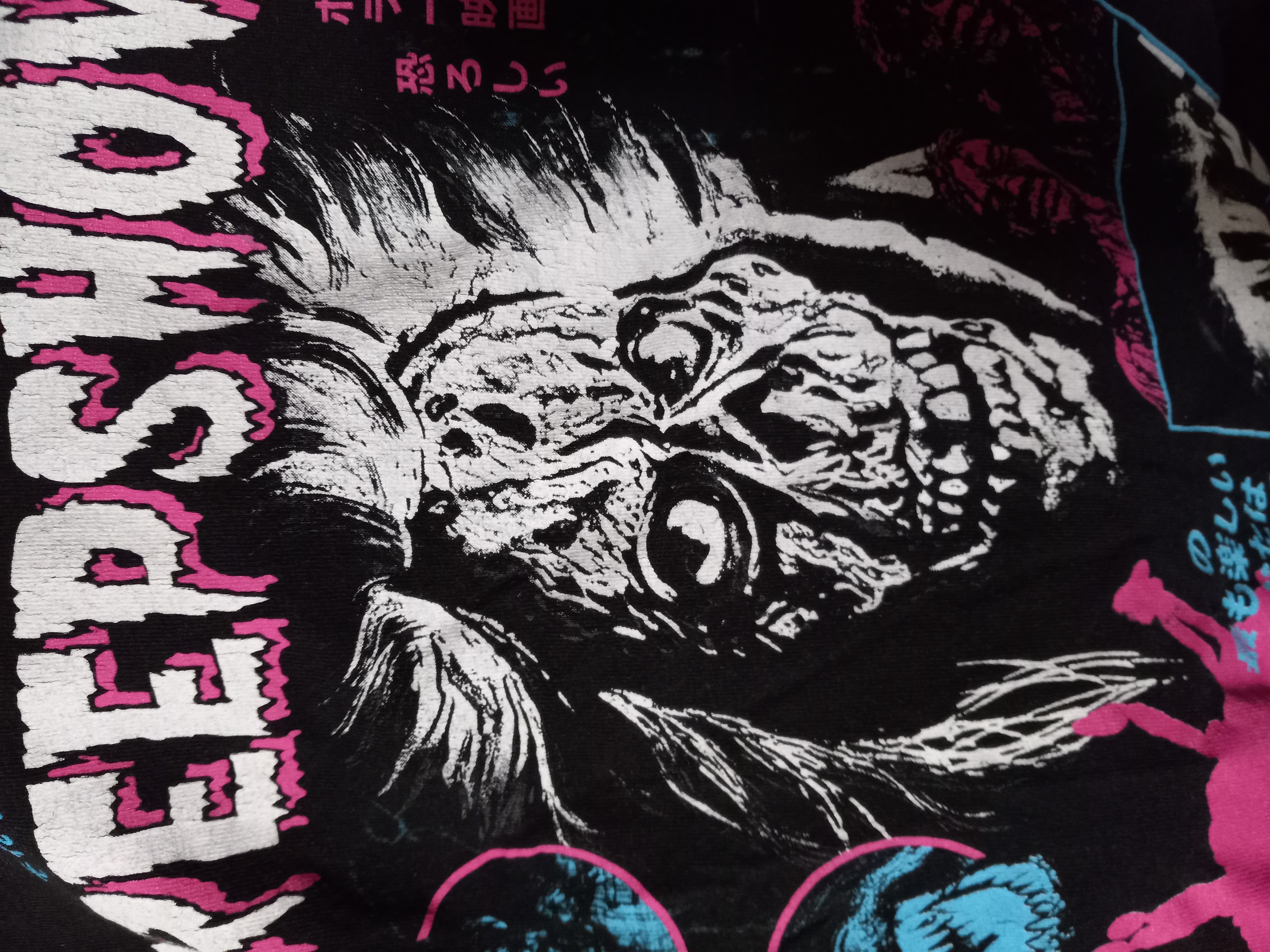 loseup shot of a black tshirt featuring a graphic design. the central element is a large, stylized skull with a zombielike appearance, depicted in shades of white and black, surrounded by pink and blue accents. the top features the word "keep show" in a stylized pink font. other design elements include japanese text and additional smaller graphics on the bottom and sides. the overall style is reminiscent of horror movie posters or punk rock aesthetics.