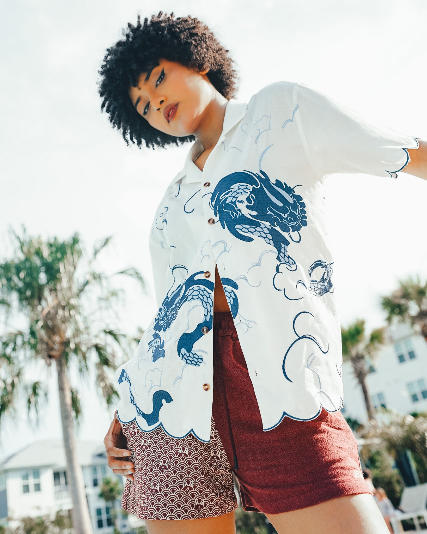 a lowangle, medium shot features a woman with dark, curly hair, wearing a white, buttondown shirt with a blue dragon and cloud print. she also wears burgundy shorts on one side and patterned shorts on the other. she is standing outdoors on a sunny day, with palm trees and buildings visible in the background. the shot emphasizes her casual summer style and confident pose looking directly at the camera.