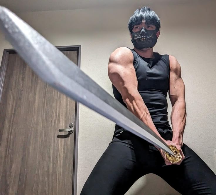 a muscular man wearing a black tank top, black leggings, and a black mask is holding a katana and posing indoors.