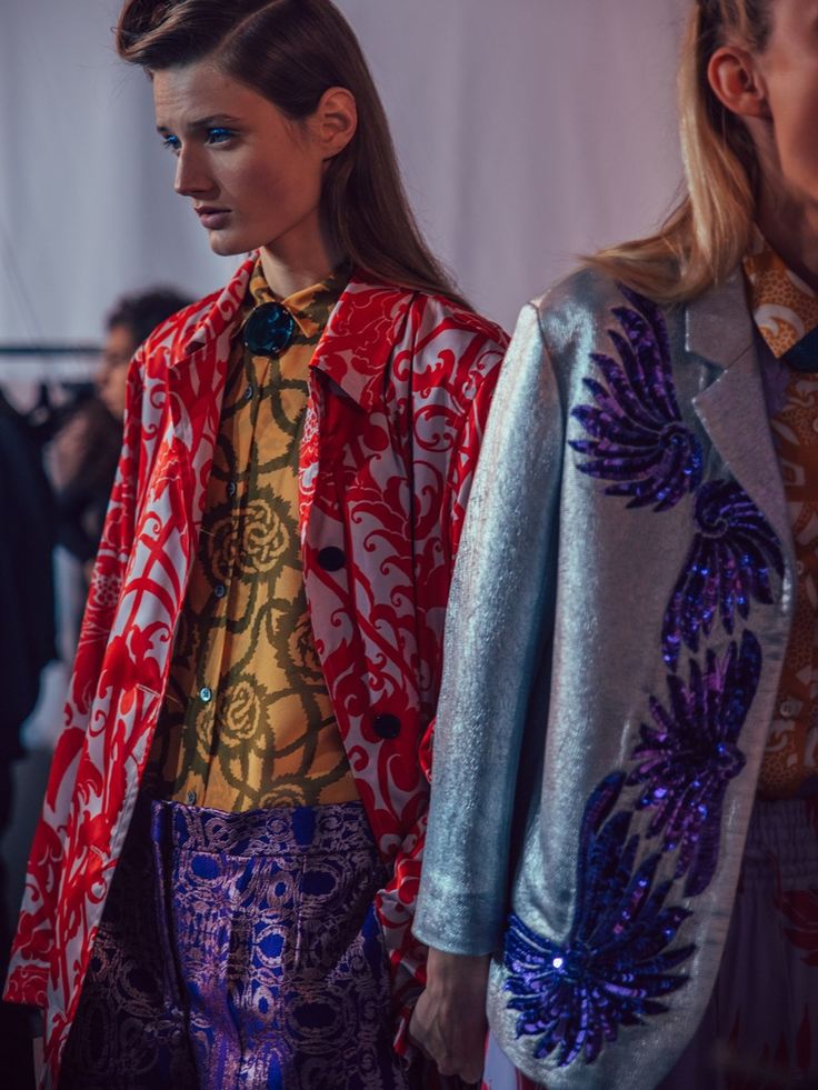 wo models are standing close together, showcasing their outfits during what seems to be a fashion show backstage. the model on the left is wearing a red and white patterned coat over a yellow patterned shirt and purple pants with an intricate pattern. her hair is styled in a sleek updo. the model on the right wears a light blue jacket adorned with purple sequins, creating a floral design. her blonde hair is partially down.  the overall ambiance suggests a fashion photography setting, focused on the details of their clothing.