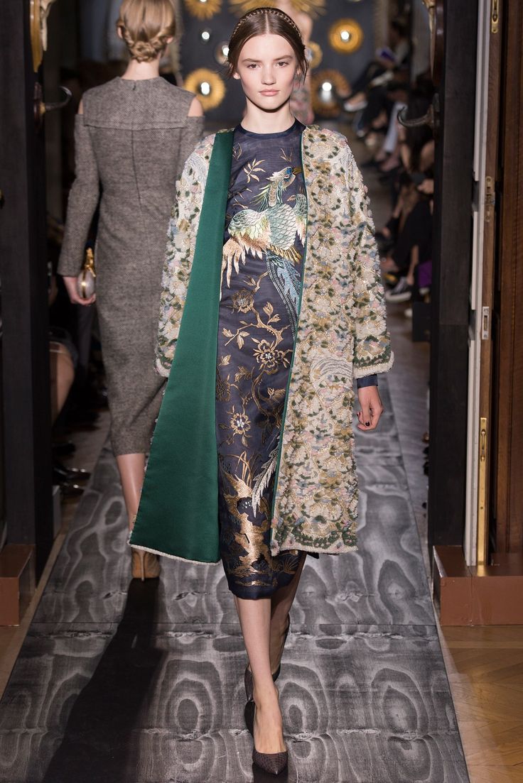 a model walks down a runway wearing a stunning haute couture ensemble. the outfit consists of an embroidered midi dress featuring peacocks and floral patterns in gold and dark colors, layered with an elegant, long green coat adorned with delicate embellishments. the model is wearing her hair neatly tied back, and she carries herself with poise and confidence. the background hints at a luxurious setting, common in fashion shows. the floor is detailed and adds to the overall opulent feel.