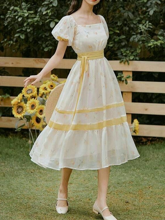 a young woman in a white and yellow floral dress is standing in a garden. the dress is midi length with puff sleeves, a square neck, and a tie waist. she is wearing white shoes and has her hair down. there are sunflowers in the background. the photo is taken from eye level in natural daylight.