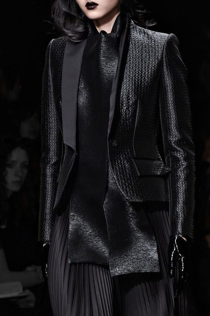 a woman is posing in a black dress and jacket with a serious expression. she is wearing a black satin dress with a pleated skirt and a black blazer with a unique textured pattern. her look is elegant and sleek. the background is a dark, blurry backdrop which highlights her outfit. the dark color palette creates a mysterious and chic vibe.