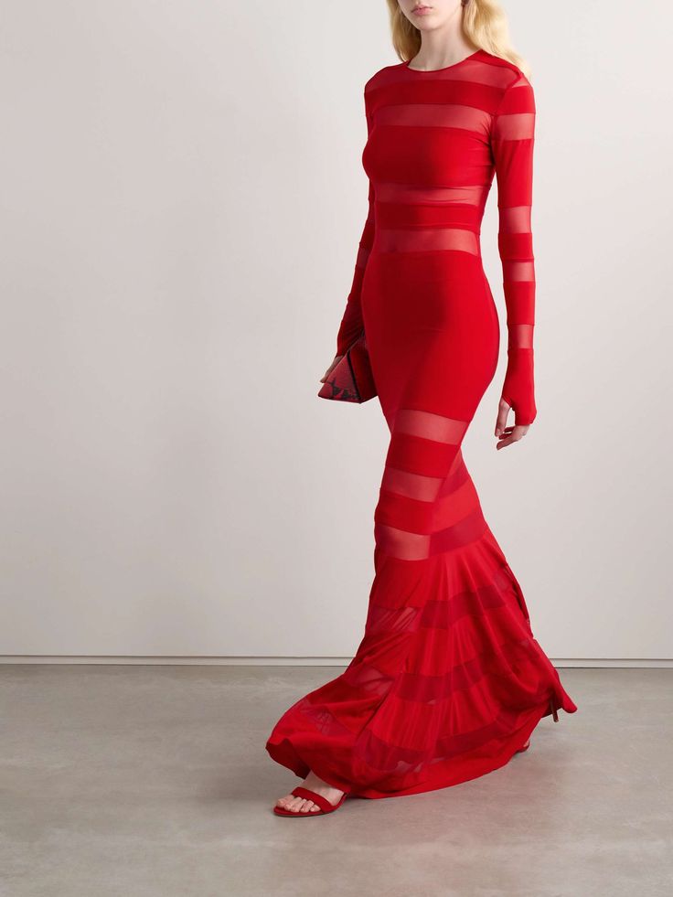 a woman is wearing a long red dress with sheer horizontal stripes. she is posing in a studio with a white background. her hair is blonde and flowing, and she is wearing red shoes. she has a red purse in her left hand.