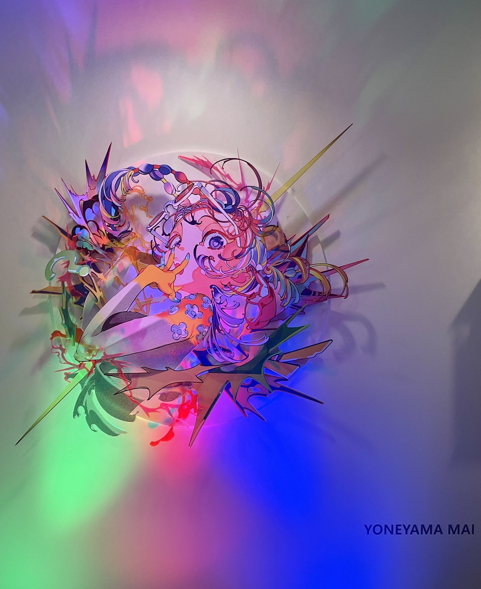 this is an artwork on display in an art gallery. it is an illustration of a woman with pink hair and a colorful outfit, done in a graphic and abstract style. the artwork is illuminated by spotlights that cast colorful shadows on the wall. the perspective of the image is from the bottom up. the overall style of the artwork is reminiscent of japanese anime and manga.