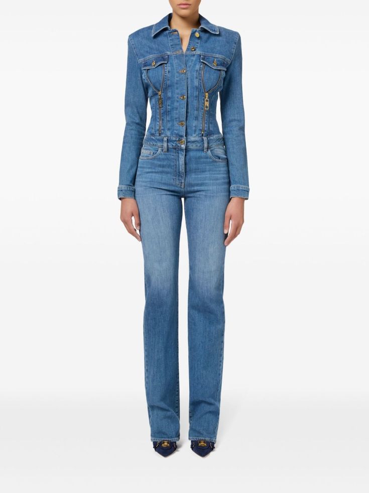 a woman is posing in a light blue denim jumpsuit. the jumpsuit has gold zippers and a buttonup jacket style top. she is wearing navy blue shoes. the image is taken in front of a white background.