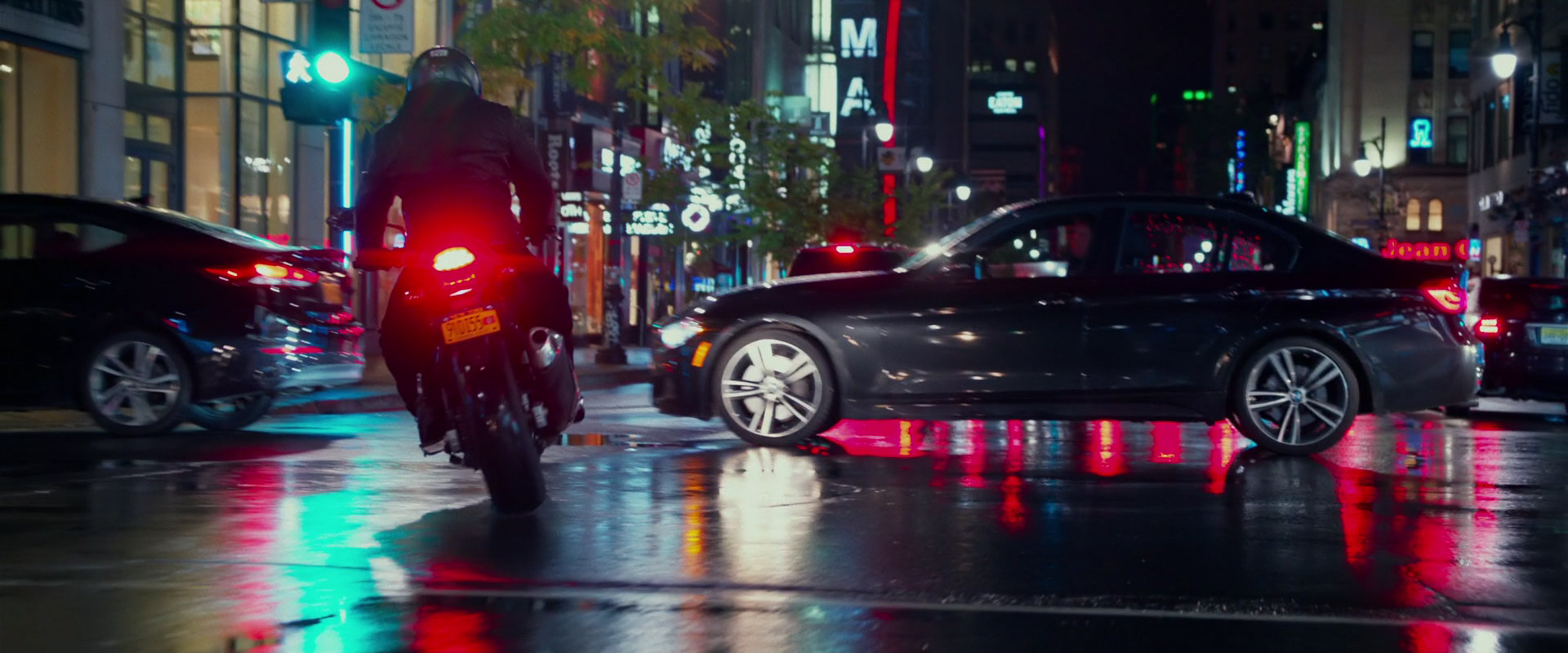 a motorcyclist in black helmet and jacket drives a black motorcycle down a city street at night. the street is wet and reflects the colorful lights from the buildings and cars. a black bmw sedan is visible to the right of the motorcycle. the scene is shot from a low angle behind the motorcycle.