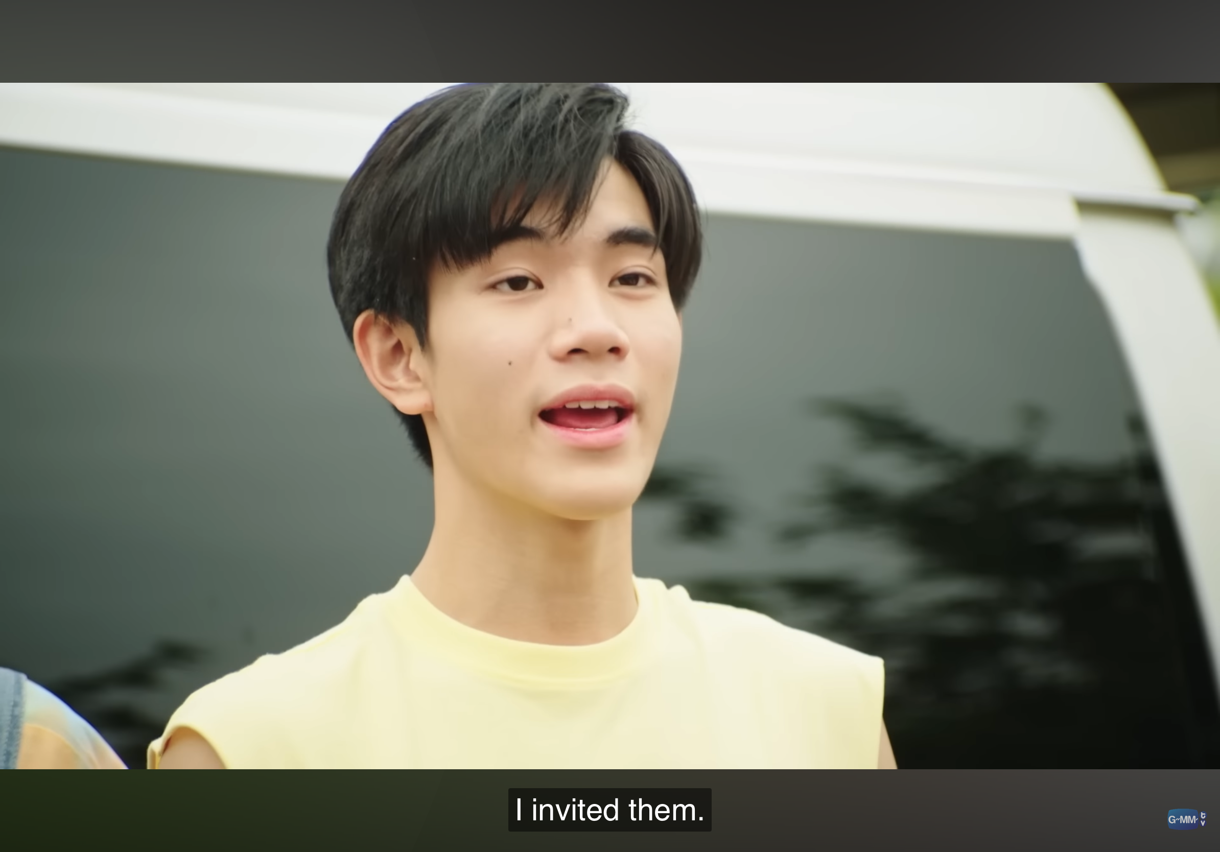 a young asian man with short black hair and wearing a yellow tshirt is talking while looking at the camera. the background is out of focus and suggests a field with grass and a vehicle. the overall lighting is bright and warm, likely from natural sunlight.  the image appears to be a still frame from a video or film, as indicated by the "i invited them." caption and the gmmtv logo in the bottom right corner.