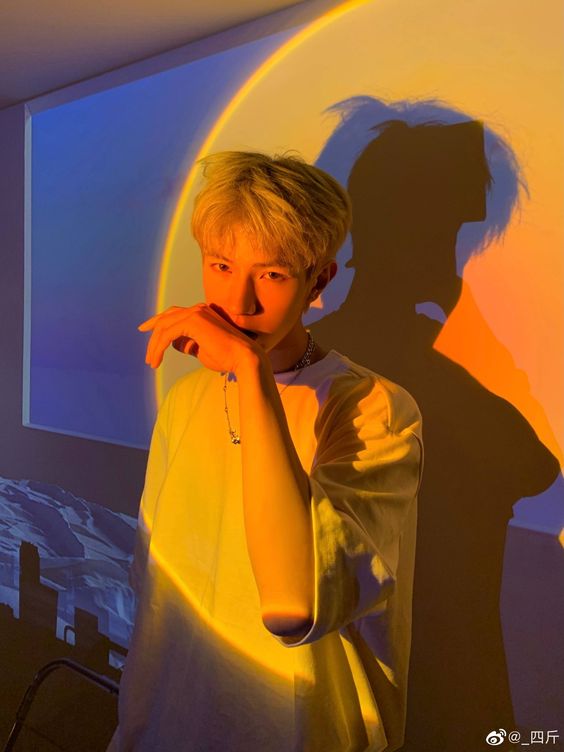 a young asian man with bleached blonde hair poses for a selfie in front of a wall projection of a sunset and rainbow. the colorful light casts a dramatic shadow of him onto the wall. he's wearing a yellow tshirt and has a silver necklace. he holds his hand near his mouth, adding to the artistic and posed nature of the picture.