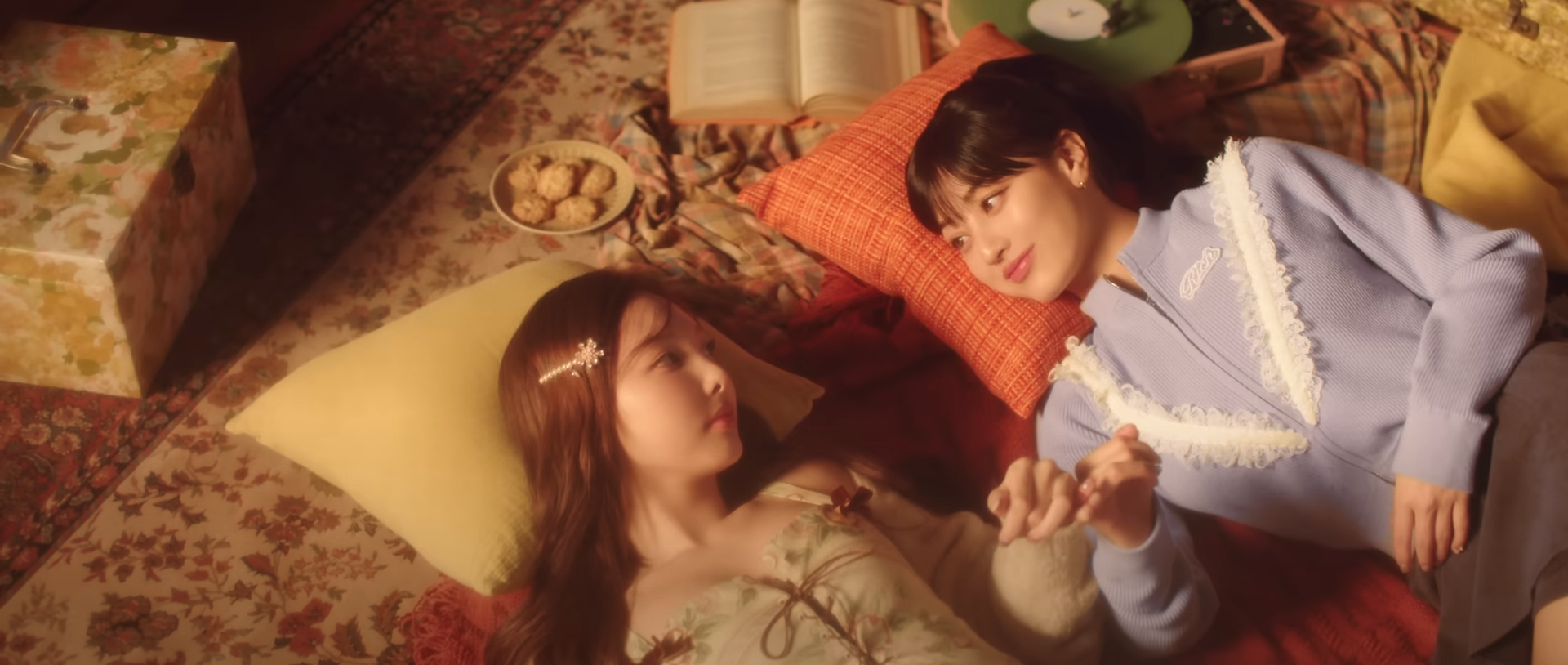 two young women lie on a vintagestyle floral rug, their heads resting on yellow and orange pillows. they are holding hands and looking at each other. the image is bathed in warm, soft light, creating a cozy and intimate atmosphere. surrounding them are objects that contribute to the retro aesthetic, including books, a record player with a green vinyl record, and a floral patterned box. the women's clothing also reflects a 70s vibe.