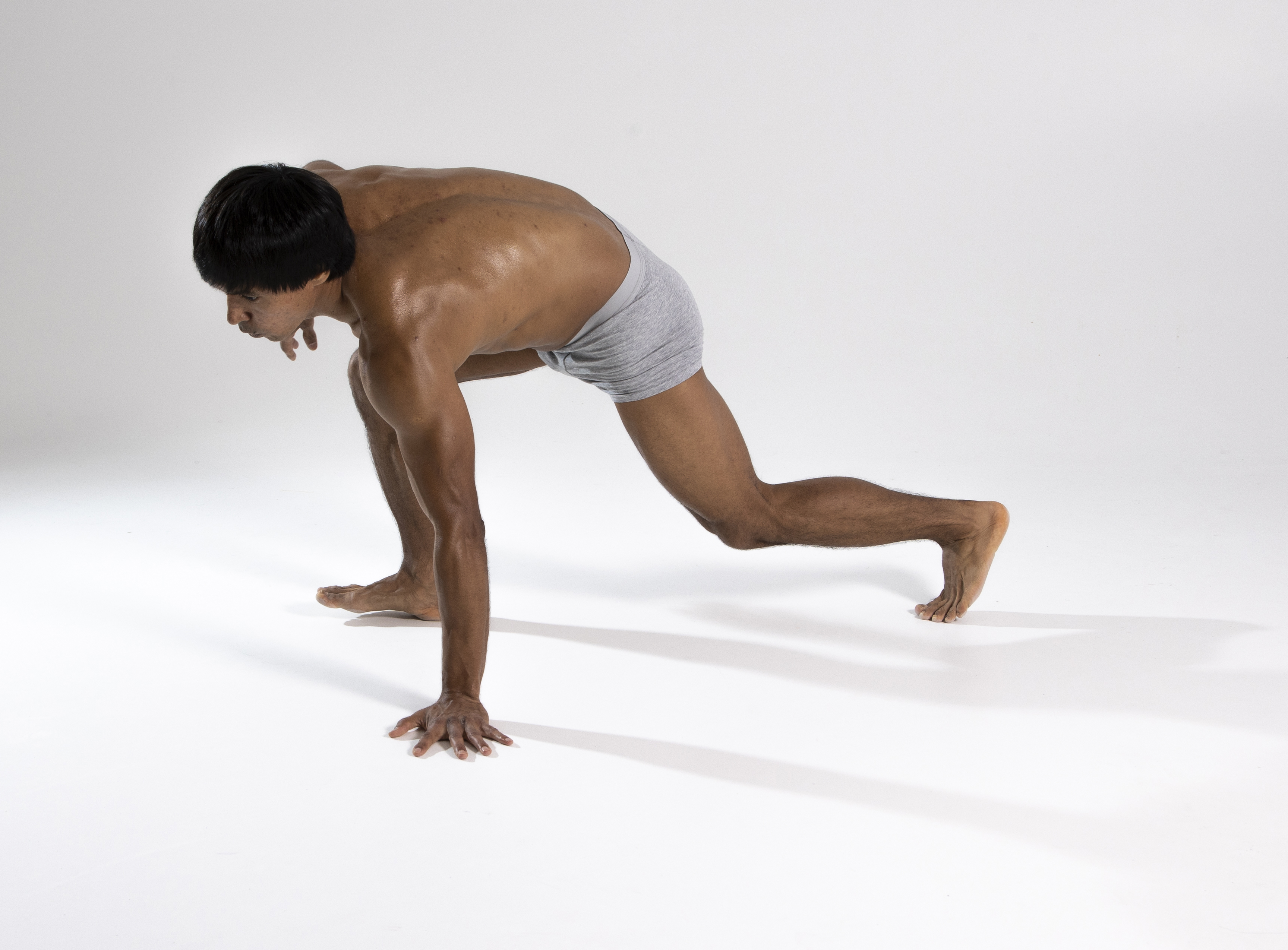 a barefoot, athletic man wearing grey underwear is doing a stretching exercise. he is in a low lunge position with both hands on the white floor. he is in a studio with a white background. his torso is bare.