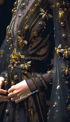 a woman is wearing a black, sheer dress with gold embellishments and intricate details. she is standing with her hands clasped in front of her, showing off the intricate beading and jewelry on the dress. the dress has a vintage, glamorous aesthetic and is adorned with sparkles and shine. the overall look is elegant and sophisticated.