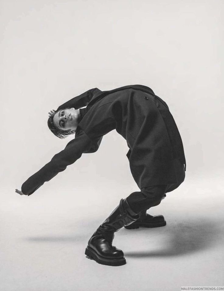 the image is a black and white fashion photograph of a male model. he is wearing a long, darkcolored coat and high black boots. the model is dramatically posed in a curved, almost backbending position, showcasing the clothing.  the background is a simple, light grey, keeping the focus on the model and his attire. the overall style is minimalist and contemporary, with an artistic and elegant feel. the image appears to be from a fashion editorial or campaign.