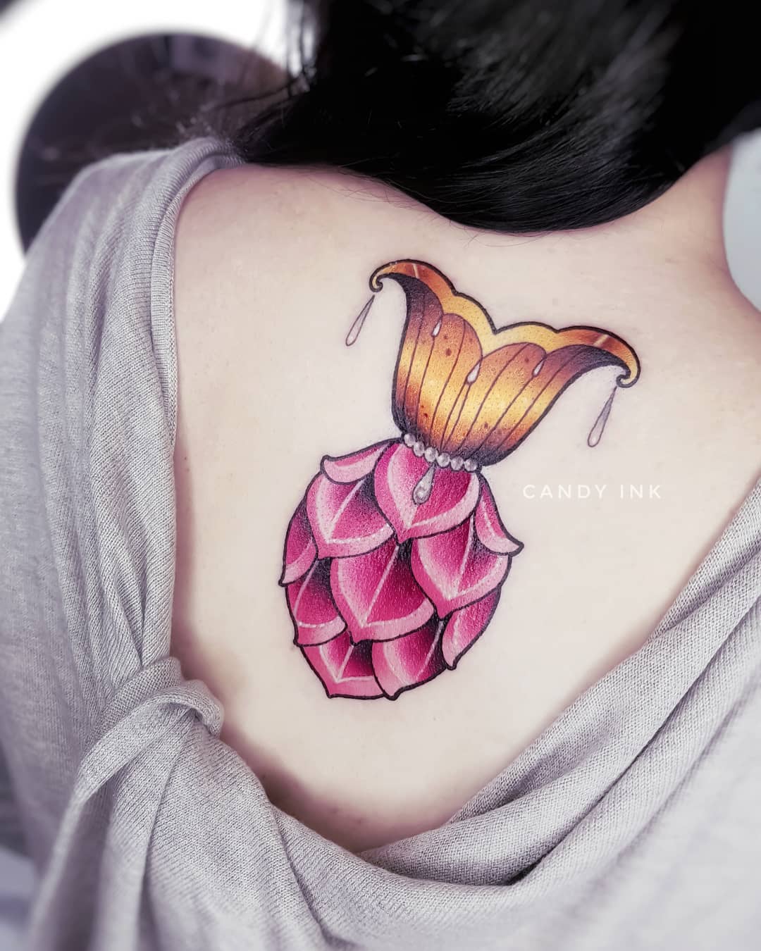 this is an image of a pink and gold jeweled flower tattoo on a woman's back, just below her shoulder. the image is taken from behind the woman and shows the tattoo in full. the background is white.