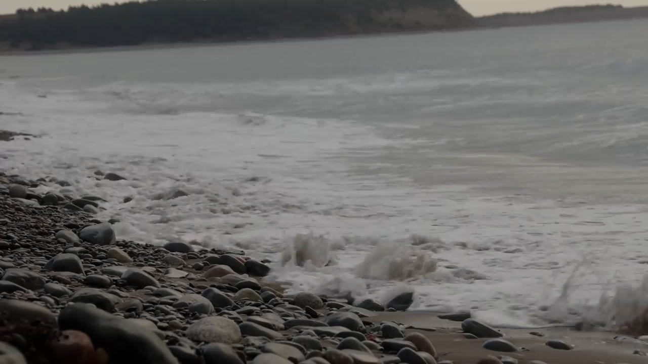 the image shows a rocky beach with waves crashing on the shore. the sea is choppy and the sky is overcast. the beach is covered in small, smooth rocks.