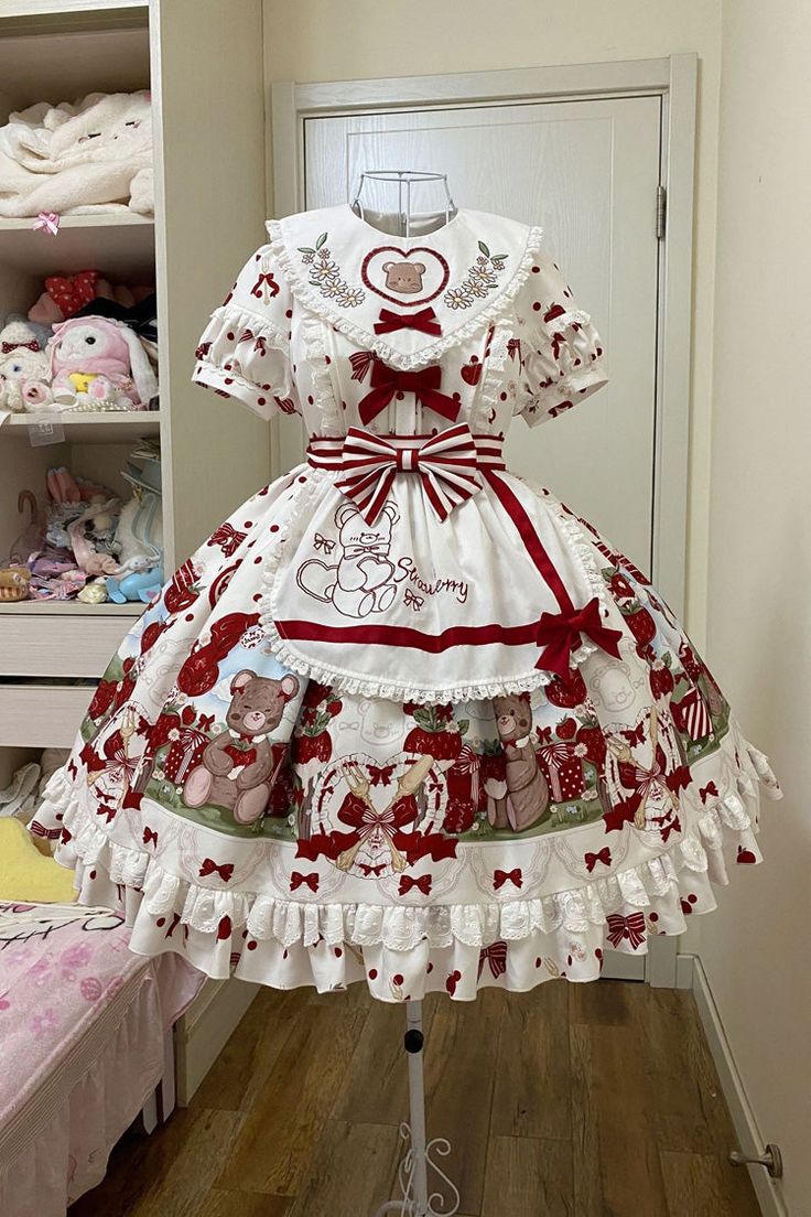 the image shows a lolita fashion dress displayed on a mannequin. the dress is white with red polka dots and features embroidery of teddy bears and strawberries. the dress has a square neckline and short, puffed sleeves, and a full, floorlength skirt with multiple layers of ruffles and lace. there is a red ribbon tied around the waist and a matching apron with more teddy bear embroidery.  the dress is hanging in a room with a white closet and a pink blanket visible in the background.