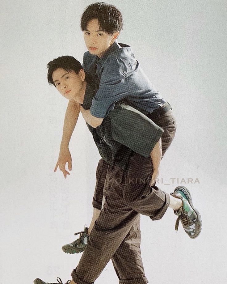 two young men are having fun while posing for a photoshoot. the man on the bottom is carrying the other man on his back as they both smile and look at the camera. the man on the bottom is wearing brown pants and a black shirt while the man on top is wearing denim jeans, a blue shirt and a black jacket. they both are wearing sneakers, and the man on the bottom is wearing black sneakers with brown laces while the man on top is wearing green and black sneakers. the background is a white wall.  the photo has a casual and fun vibe.