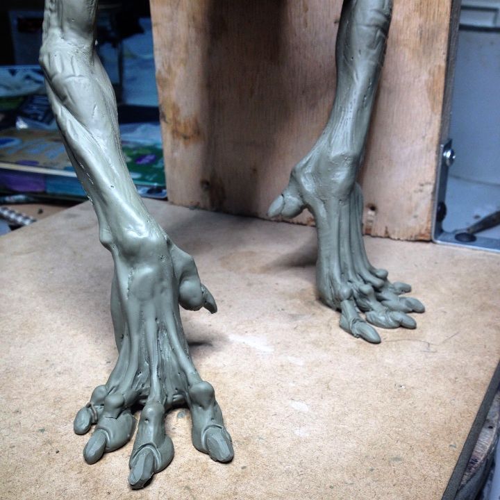 the image shows a closeup of a gray clay sculpture featuring two thin legs and feet. the feet are large and have elongated toes. the sculpture appears to be unfinished and is set on a wooden surface. a blurred background includes hints of other materials and tools, indicating a studio or workshop environment.