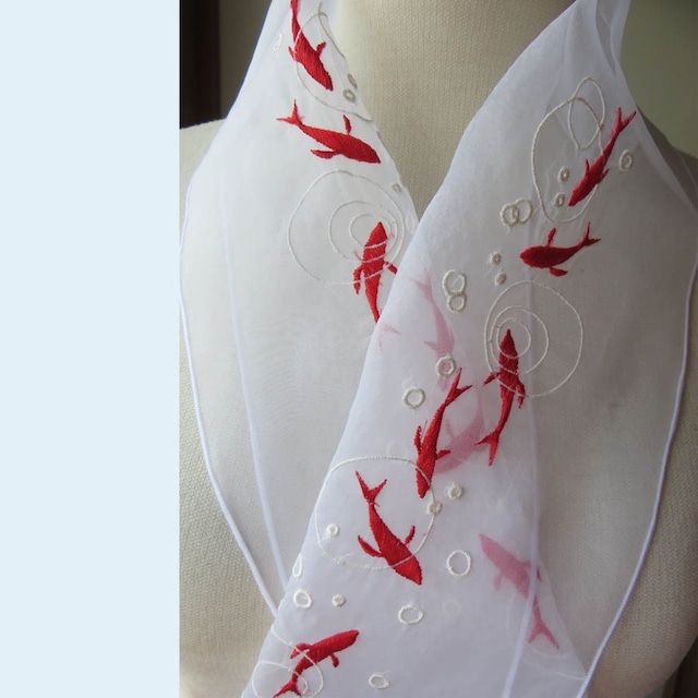 a closeup image showcasing a white, sheer scarf with delicate embroidery. the embroidery features red fish and white bubble designs. the scarf appears to be artfully draped, suggesting it might be displayed on a mannequin or model. the overall impression is one of handcrafted elegance and aquatic inspiration.