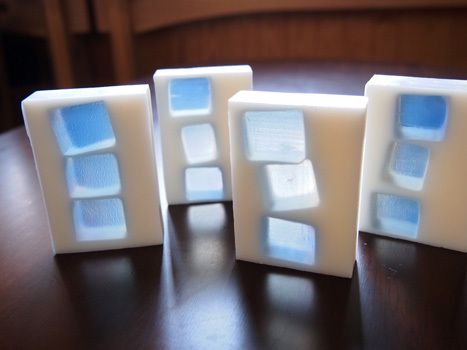 four white soap bars are stacked on top of each other. the soap bars have blue designs on them that look like they are made of glass or plastic. the soap bars are on a dark wooden surface and have shadows below them. the blue designs on the soap bars are very intricate and have a lot of detail. the soap bars look very clean and new.