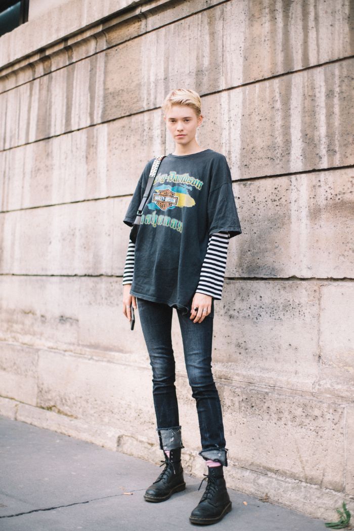 a stylish young woman with short blonde hair stands against a stone wall in a casual yet edgy outfit. she is wearing a black harleydavidson graphic tshirt layered over a black and white striped longsleeved shirt, dark blue skinny jeans cuffed at the ankles, and black doc martens boots. she carries a crossbody bag. the overall style is grungeinspired and fashionable.  the setting appears to be an urban environment.