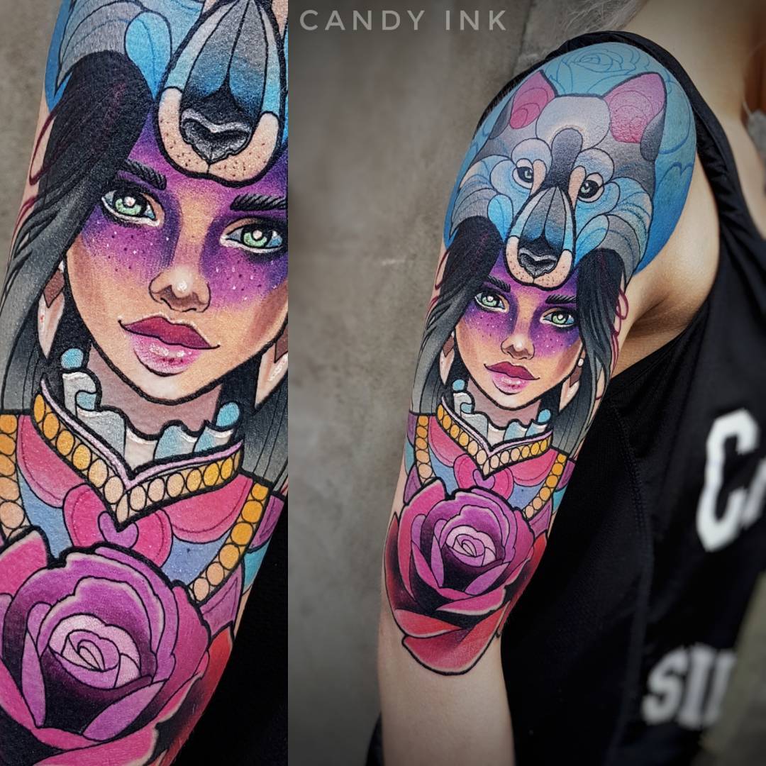 a person's left arm with a colorful tattoo of a woman with a wolf headdress. there is a large rose tattoo on her arm and she has purple and blue eye shadow.