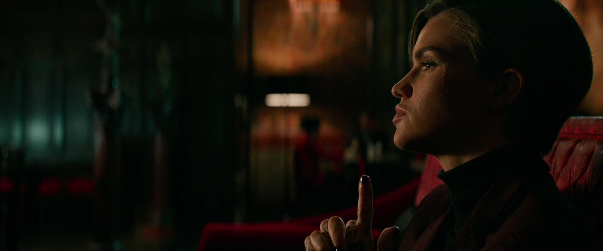 a woman with short hair is sitting on a red couch in a dark room, looking off to the side. her face is serious and her finger is raised to her lips, as if she is thinking about something.