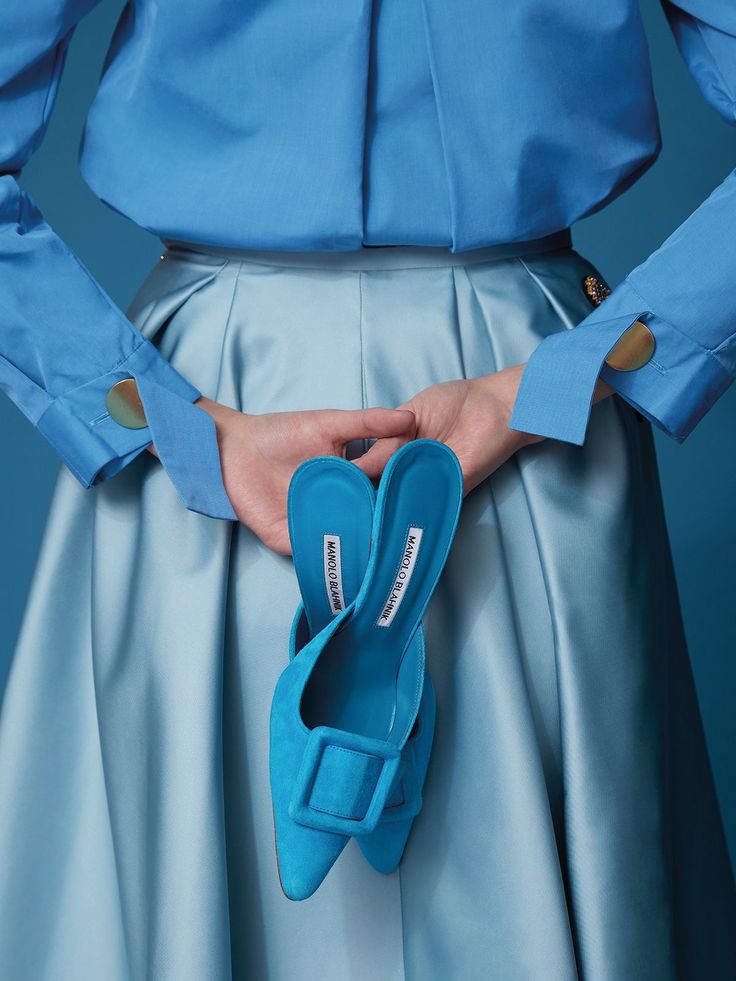the image shows a woman from the midtorso down, wearing a light blue, longsleeved shirt tucked into a matching light blue, pleated midilength skirt.  her hands are behind her back, holding a pair of vibrant blue manolo blahnik pointedtoe mule shoes with a distinctive buckle detail. the overall aesthetic is monochromatic, emphasizing the different shades of blue in a highfashion context. the photography style is clean and stylish, focused on the clothing and accessories.
