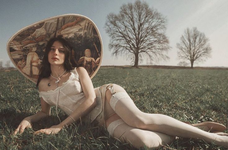 a woman is lying down on a grassy field. she is wearing lingerie, stockings, and a garter belt. she has a large, decorative hat on her head with an image resembling artwork. she is also wearing a necklace. there are trees in the background. the style of the image appears vintage.
