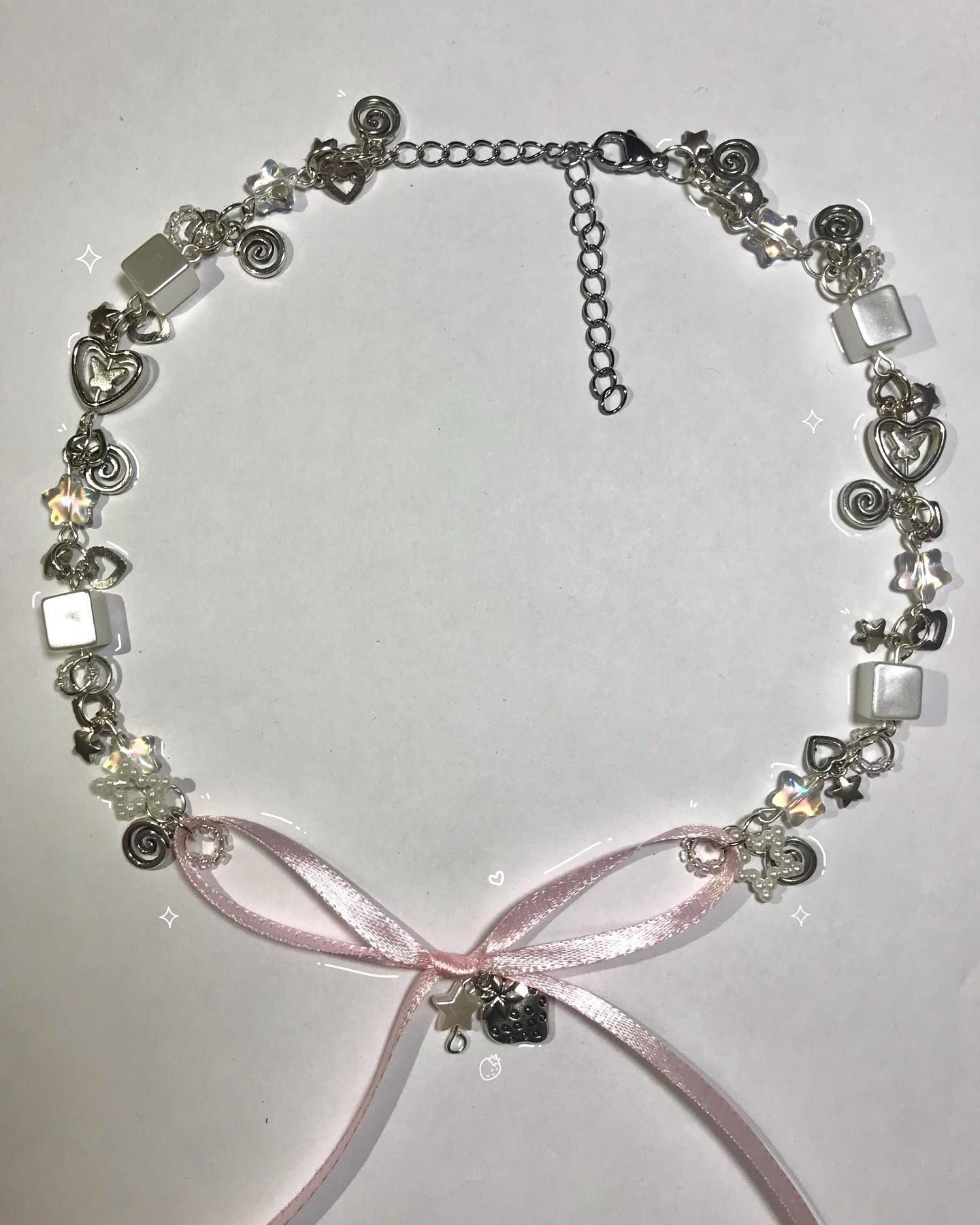 a necklace with a light pink ribbon tied at the bottom is laid out on a white surface. the necklace is silver and features a variety of charms and beads, including hearts, stars, swirls, and square white beads. a hello kitty charm dangles from the ribbon's knot. the necklace also has an adjustable chain at the back.