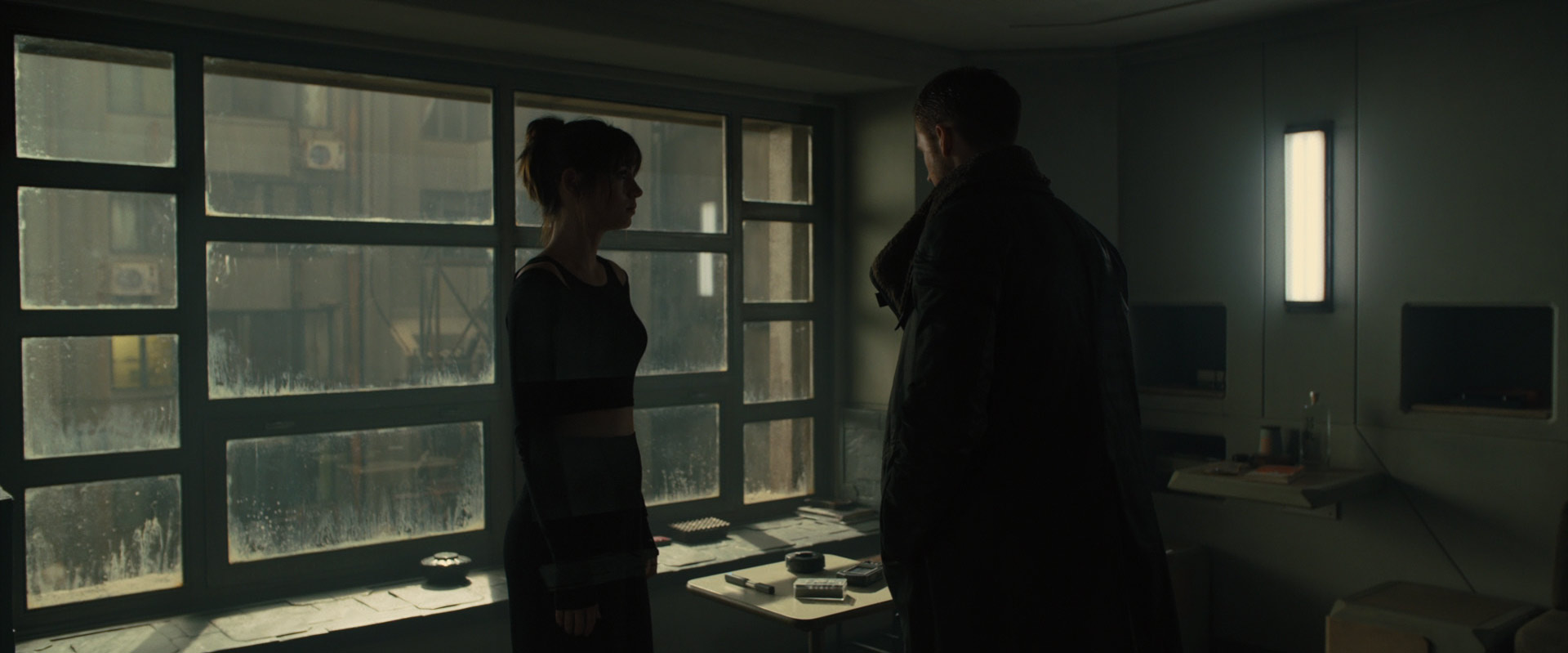 a woman and a man stand in a dark room with a large window looking out onto a city. the man is wearing a dark coat and the woman is wearing a dark top. the room is lit by a single bright light on the wall and the city lights from outside. they are both looking at each other and appear to be in conversation.