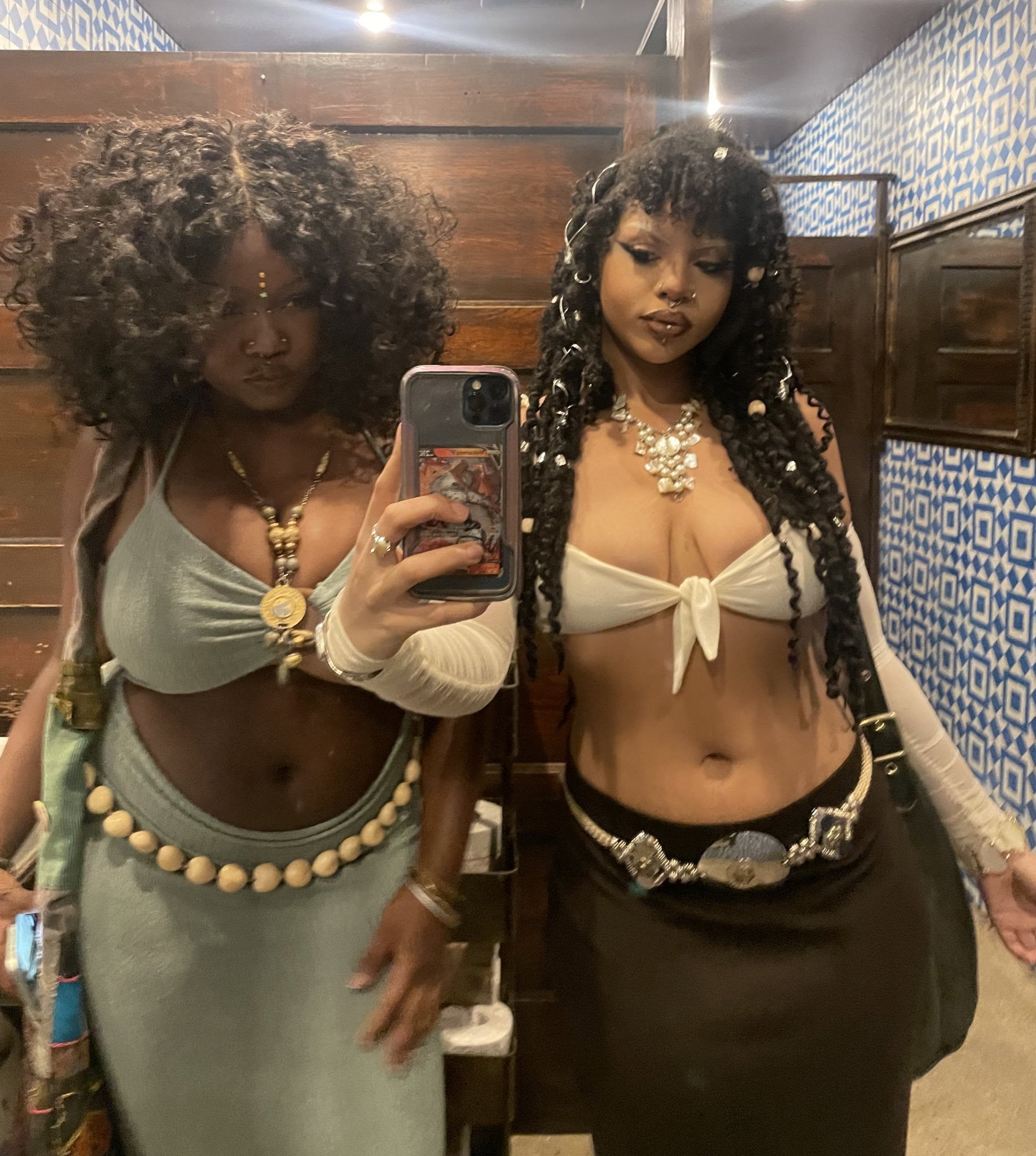 two young women, one with a crop top and skirt, and the other with a long skirt and halter top, pose in a mirror, smiling while taking a selfie. both women are wearing jewelry and have long hair, one with braids. they appear to be friends.