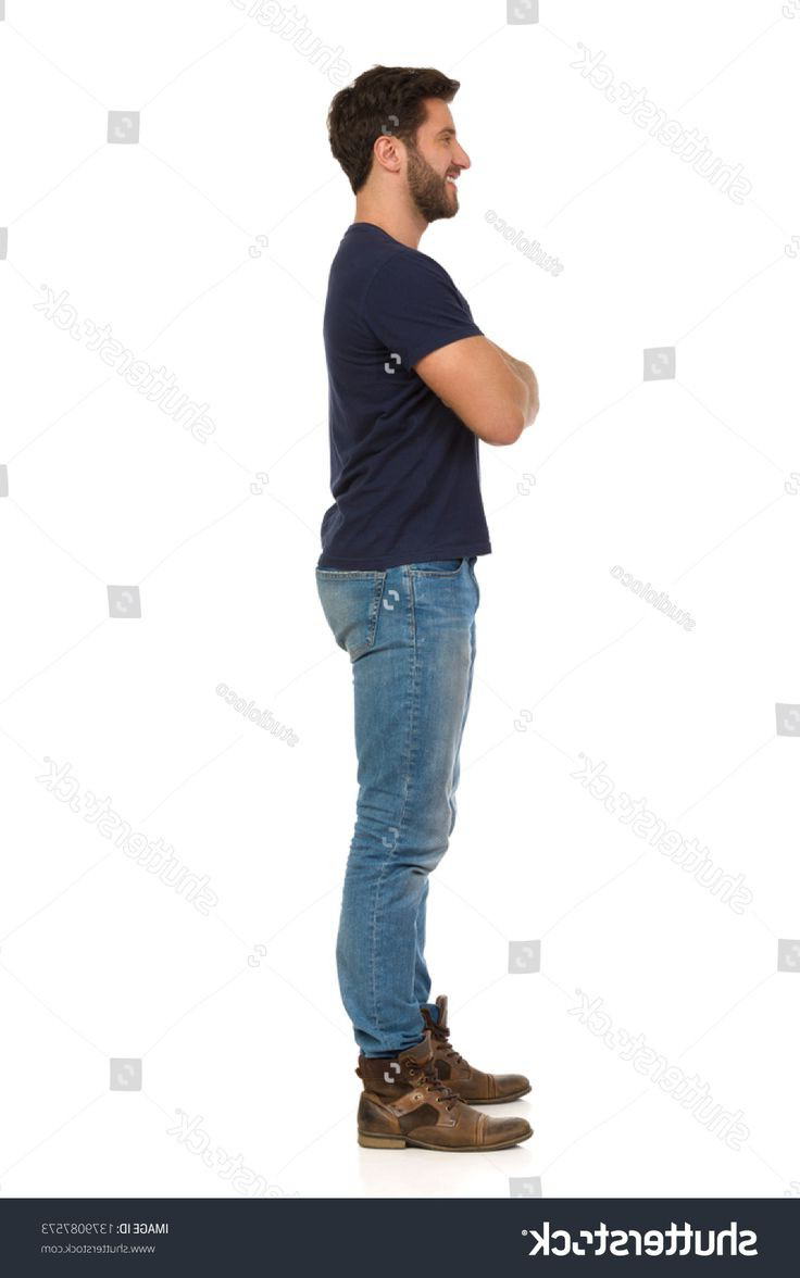 a man in a blue tshirt and jeans is standing with his arms crossed and looking to the side. he is wearing brown boots and has a slight smile on his face. he is standing in front of a white background.