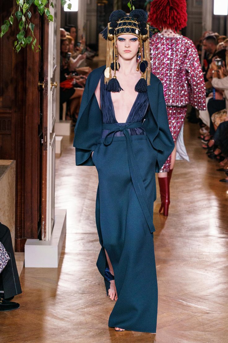 a model walks the runway in a chic, long teal dress with a plunging neckline and a dramatic train. her head is adorned with a large, elaborate gold and black hat. the dress is cinched at the waist with a gold belt and the model is wearing dark, highheeled boots. the model is walking down a wooden catwalk in front of a large audience of fashion enthusiasts.