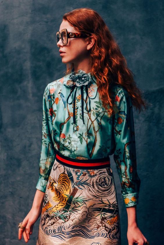 a stylish redhead model with curly hair is posed against a dark teal textured background. she is wearing a teal silk gucci shirt with a floral and bird print. the shirt is tucked into a highwaisted aline skirt featuring a tiger and wave print in shades of beige and brown.  a red and green webbed belt sits at her waist, and she accessorizes with large squareframed sunglasses. the overall style is vintageinspired and elegant. the image appears to be a professional fashion shoot, likely showcasing gucci's clothing line.