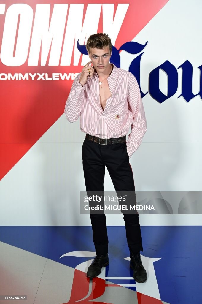 a fulllength shot of a handsome young man posing in front of a tommy hilfiger backdrop. he is wearing a light pink striped shirt, which is unbuttoned at the top, black pants, a brown belt, and black shoes. he has blond hair and is touching his ear with his right hand. the backdrop features the tommy hilfiger logo in red, white, and blue.