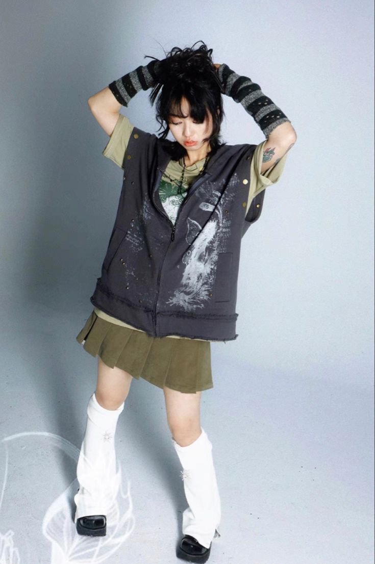 a young woman is posing in a studio against a light grey background. she is wearing a grey sleeveless hoodie with a graphic print, a green pleated skirt, white leg warmers, and black shoes. her hair is pulled back and she is wearing dark grey striped arm warmers. she is looking down.