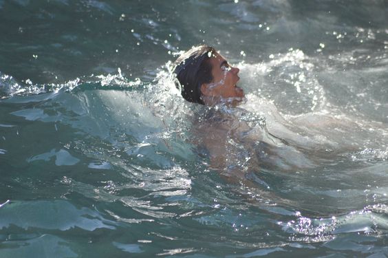 a young man is struggling to stay afloat in rough ocean water.  the sun is shining brightly, creating sparkling reflections on the choppy waves. the perspective is at eye level, giving a sense of immediacy and danger.