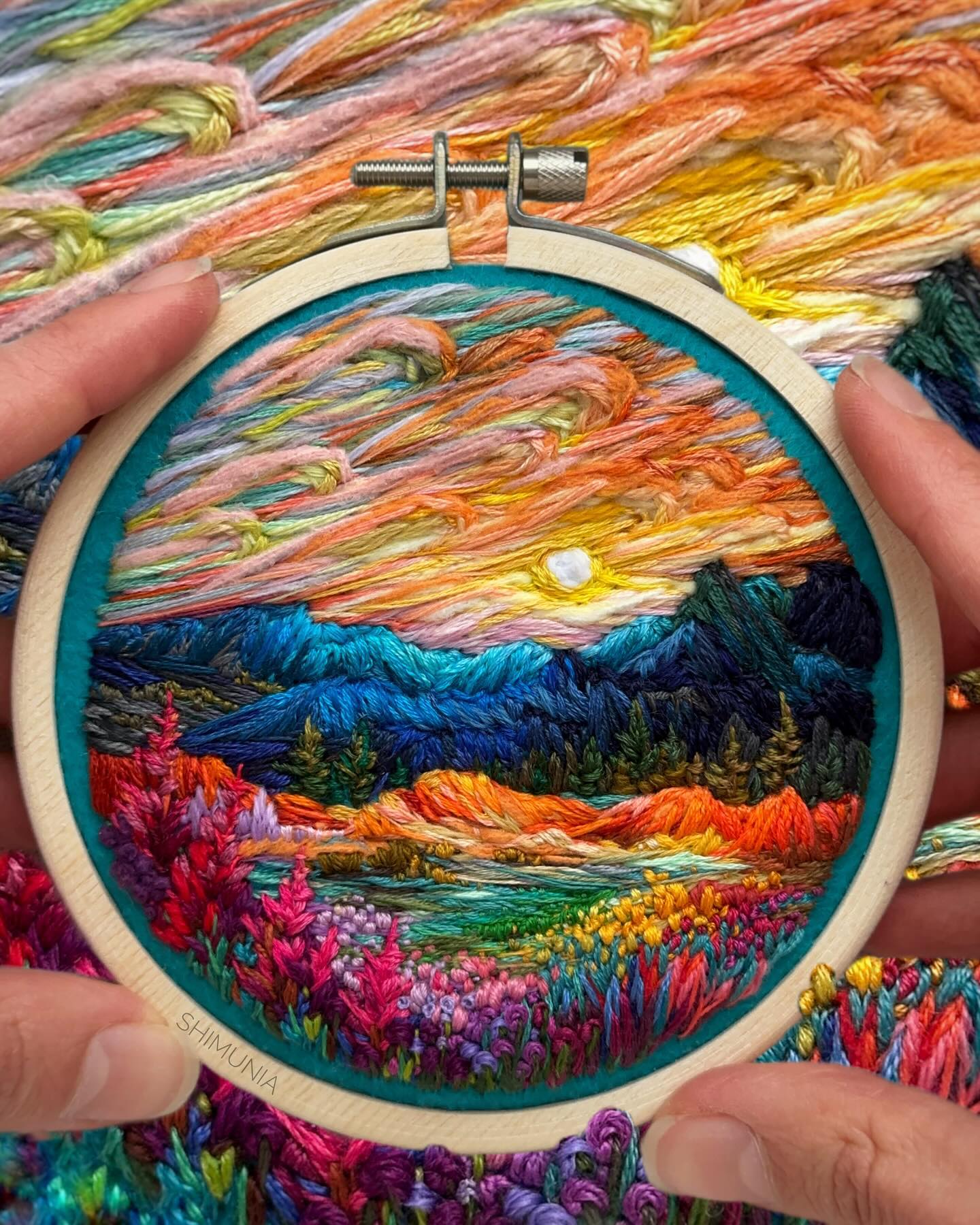the image showcases a stunning embroidery hoop art piece depicting a breathtaking sunset over a mountainous landscape. the scene is brought to life with vibrant threads capturing the warm hues of the setting sun reflecting on the peaks and illuminating the clouds. lush greenery covers the foreground, while a serene lake reflects the beauty above. the artwork is held by two hands, emphasizing its intricate details and the artist's skillful craftsmanship.