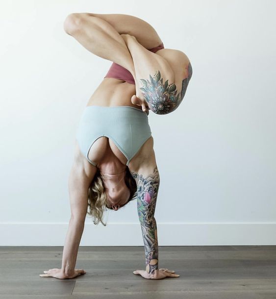 a woman with tattoos on her arms and leg is doing a yoga pose indoors. she is upside down, balancing on her hands with her legs in the air. she is wearing a blue sports bra and maroon underwear.  the background is a white wall with light brown wood flooring.